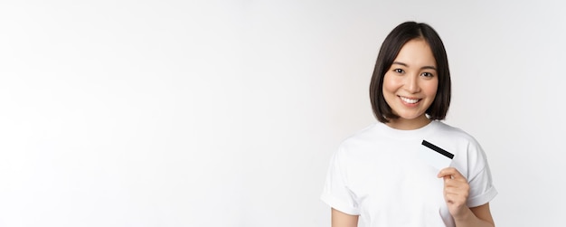 Free photo portrait of korean smiling girl bank client showing credit card with happy face standing against whi