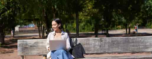 Free photo portrait of korean woman sitting with laptop in park on bench working outdoors student doing