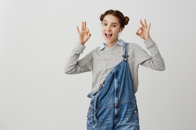 Portrait of lady in casual gesturing alright sign with both hands. Excited female customer expressing gladness after speaking with mobile operator through earpieces. Human emotions