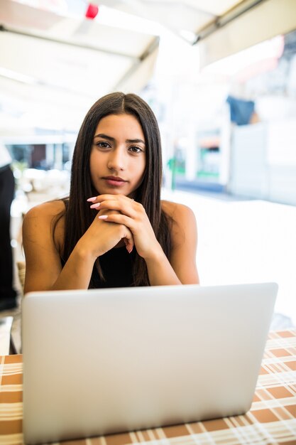 Ritratto di donna latina con gli occhiali a lavorare con il suo laptop all'aperto. affascinante donna freelance con i capelli ricci è seduta in un bar di strada e sta facendo il suo lavoro a distanza sul netbook