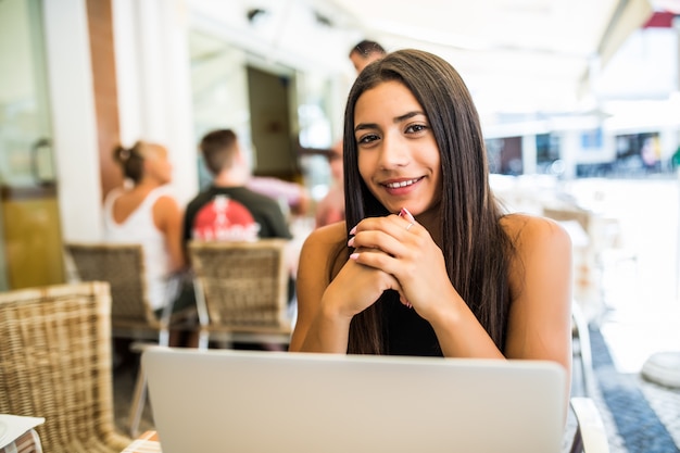 Portrait of latin woman in glasses working with her laptop outdoors. Charming freelancer woman with curly hair is sitting in street bar and doing her remote work on the netbook