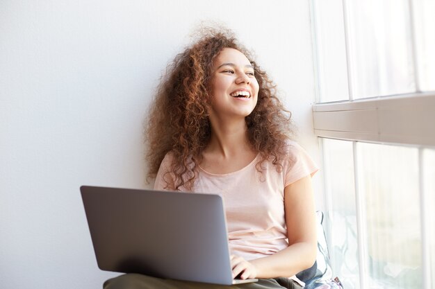 Free photo portrait of laugh young curly african american girl sitting by the window, dressed in pink t-shirt, smiling and enjoying his favorite video at freelance work.