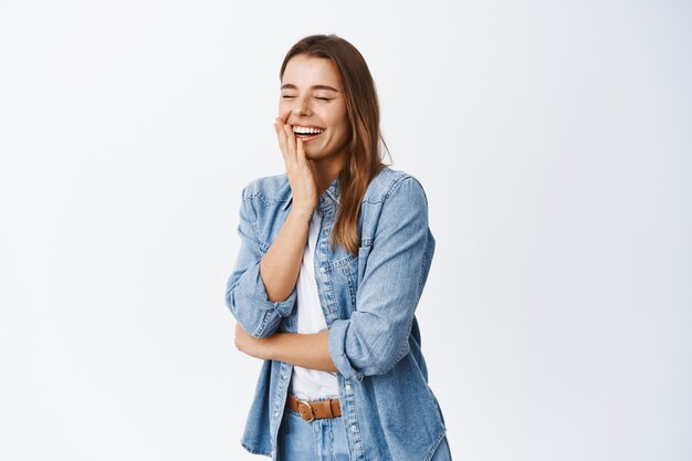 Portrait of laughing woman feeling happy, having fun, close eyes and chuckle from funny joke, standing in casual clothes against white wall