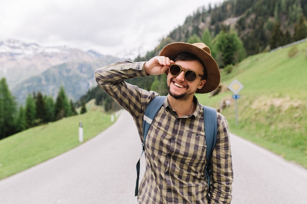 Portrait of laughing young man with beard holding sunglasses and posing on the road on Alps