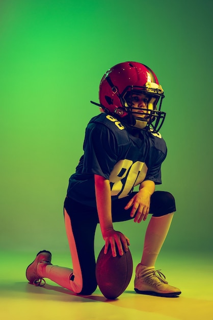 Portrait of little boy child playing training american football isolated over green background in neon