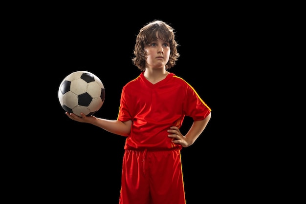 暗いスタジオの背景に分離されたサッカーボールでポーズをとる小さな男の子のサッカーサッカー選手の肖像スポーツゲームの趣味と子供の頃の概念