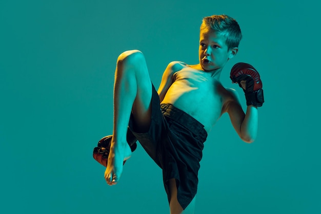Portrait of little boy kid in gloves training practicing thai boxing on green studio background in neon light