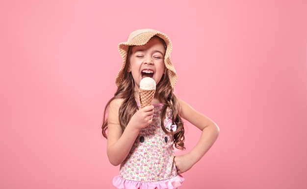 Portrait of a little cheerful girl in a summer hat with ice cream in her hands, on a colored pink background, summer concept