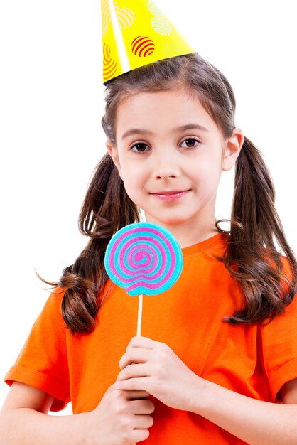 Портрет маленькой милой девочки в оранжевой футболке и партийной шляпе с цветными конфетами - изолированные на белом