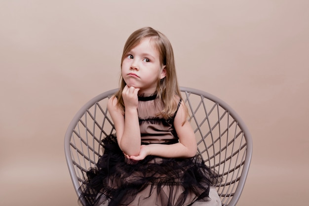 Free photo portrait of little cute girl wears black dress sitting in chair with a pensive face and posing on isolated wall, real serious motions of pretty charming girl