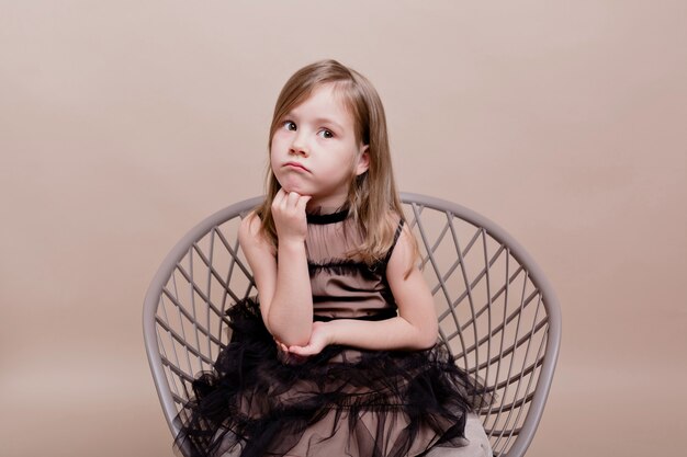 Портрет маленькой милой девочки в черном платье, сидящей в кресле с задумчивым лицом и позирующей на изолированной стене, настоящие серьезные движения довольно очаровательной девушки