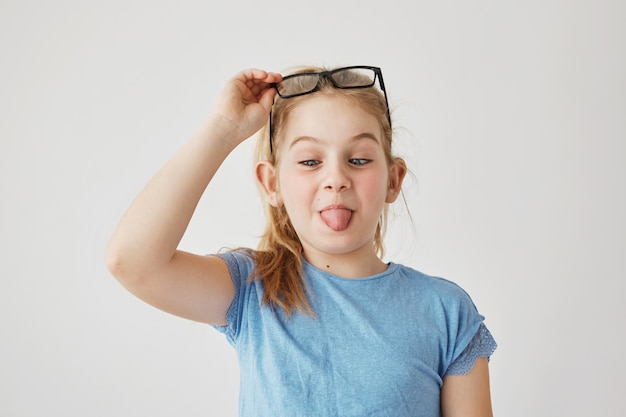 Portrait little cute miss with blue eyes and light hair in blue t-shirt funny posing with mow eyes, showing tongue and raising glasses. Copy space.