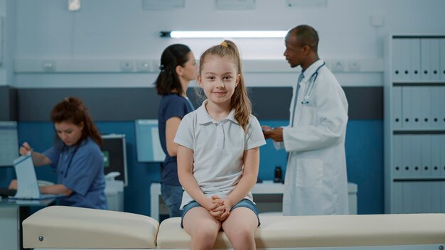 医者と一緒に医療キャビネットで健康診断を開始するのを待っているベッドの上の少女の肖像画。カメラを見て、オフィスで健康診断の訪問予約の準備をしている病気の小さな子供。