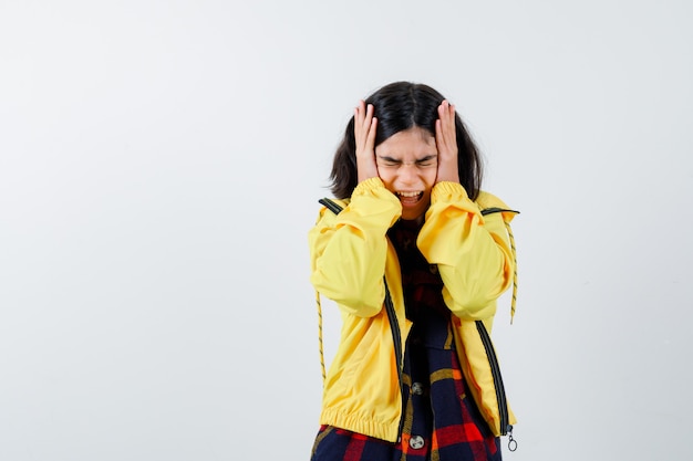 Portrait of little girl holding hands on head in checked shirt, jacket and looking confused front view