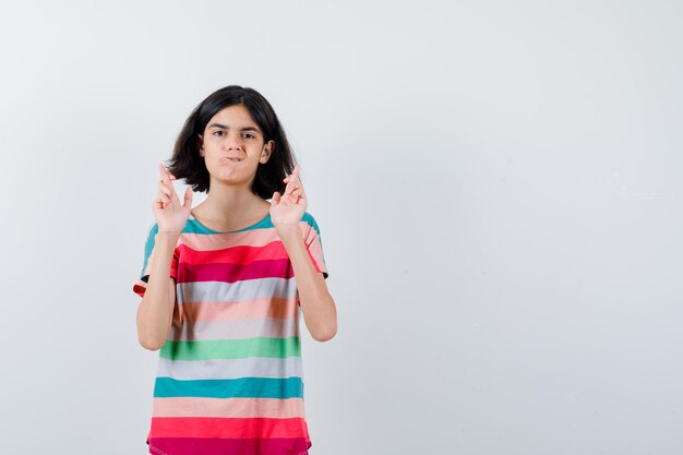 Portrait of little girl keeping fingers crossed in t-shirt and looking hopeless front view