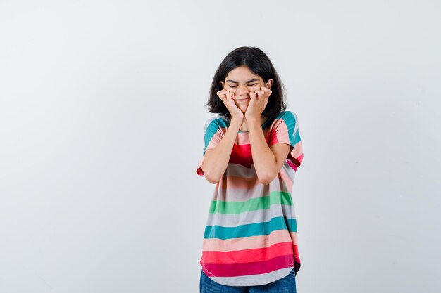 Портрет маленькой девочки, держащей руки за щеки в футболке и обиженной, вид спереди
