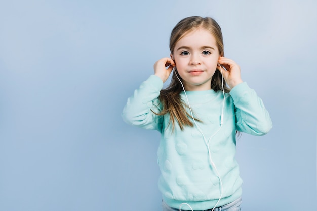 Portrait of a little girl putting earphones on her ears against blue background