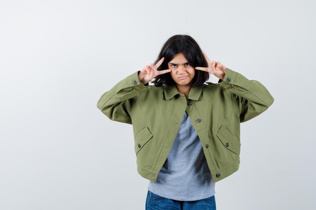 Portrait of little girl showing victory sign in coat, t-shirt, jeans and looking lucky front view