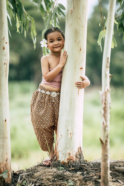 태국 전통 드레스에 어린 소녀의 초상화와 그녀의 귀에 흰 꽃을 넣어, 서서 나무의 트렁크를 포용, 미소, 복사 공간