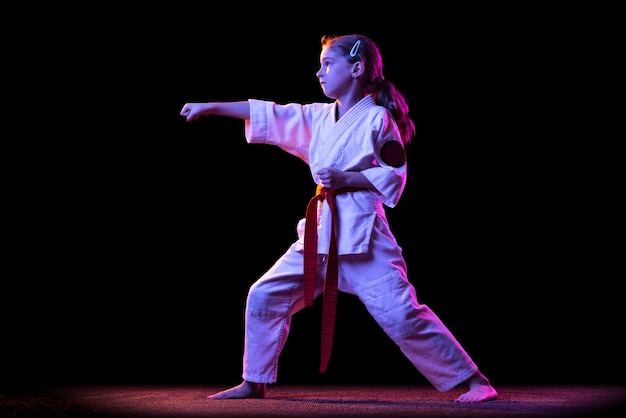 Portrait of little girl in white kimono training martial art isolated on black in neon