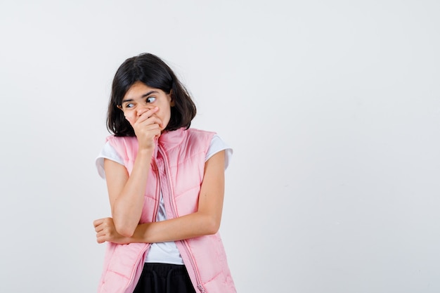 Portrait of a little girl in white t-shirt and puffer vest covering her mouth