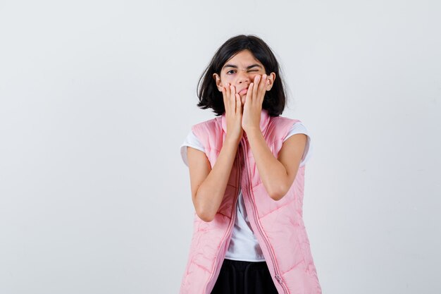 Portrait of a little girl in white t-shirt and puffer vest holding hands on cheeks