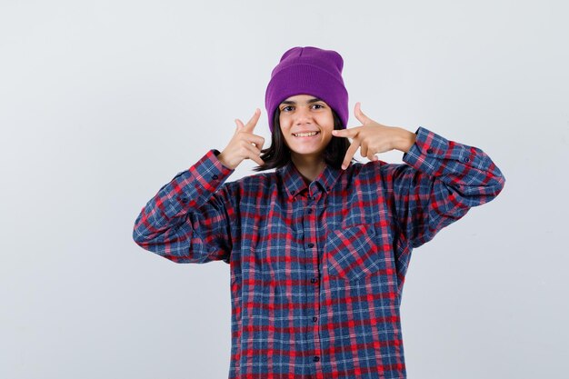 Portrait of little woman pointing aside in checkered shirt and beanie looking happy