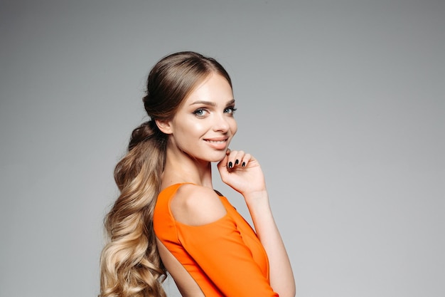 Portrait of long haired blonde woman wearing in orange outfit smiling and touching her face by fingers Girl with perfect skin and professional make up posing at grey studio background