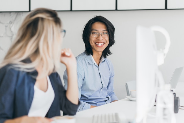 Portrait of long-haired laughing asian man with blonde woman. Pleased chinese office worker in blue shirt joking with female colleague at workplace.