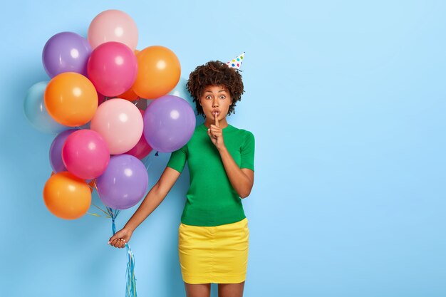 Portrait of lovely Afro American woman makes silence gesture, holds bunch of colored balloons, wears green t shirt and yellow skirt, tells secret