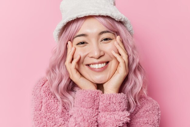 Portrait of lovely Asian woman keeps hands on cheeks smiles toothily has dyed pink hair wears winter panama and coat isolated over rosy background expresses positive emotion listens you carefully