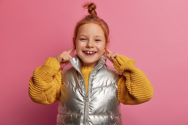 Portrait of lovely cheerful ginger girl plugs ears, has sincere smile, wears knitted sweater, vest, poses against pink pastel wall, has toothy smile, avoids listening loud music at party