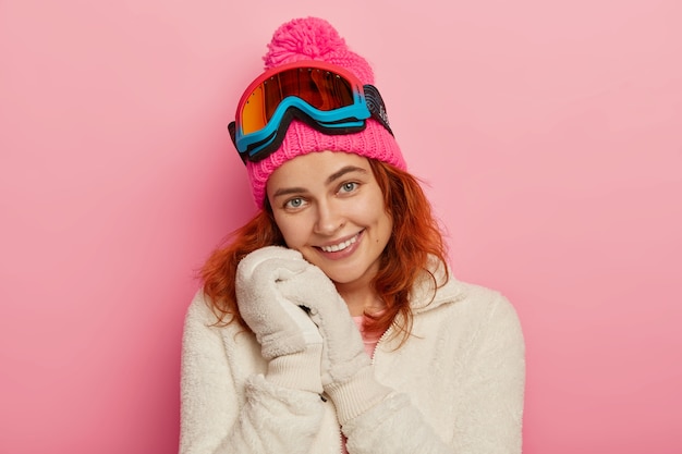 Portrait of lovely cheerful redhead woman wears soft mittens, winter white jumper,pink hat with pompon and snowboarding goggles, smiles tenderly, isolated over pink background.