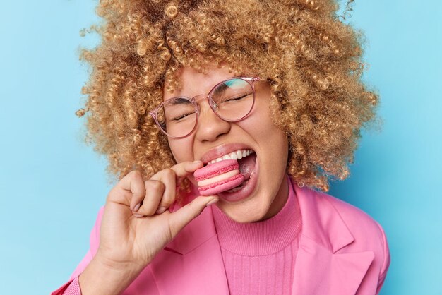 Portrait of lovely curly haired woman eats macaroon biscuit has sweet tooth enjoys unhealthy food wears optical spectacles pink jacket isolated over blue background Favorite dessert Sweetness