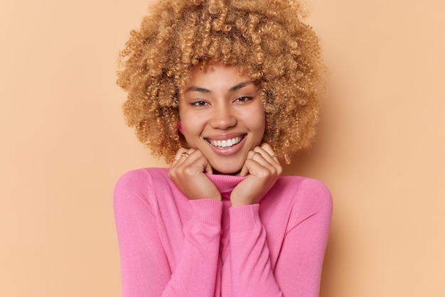 Free photo portrait of lovely curly haired young woman smiles gently keeps hands on collar of pink jumper feels glad looks directly at camera isolated over brown background pleasant human emotions concept