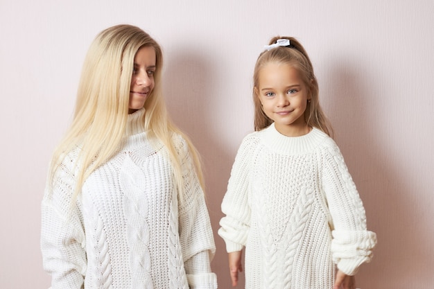 Portrait of lovely cute little girl wearing knitted jumper and ribbon in hair smiling while spending nice time with her caring loving mother who is looking at daughter with love and tenderness
