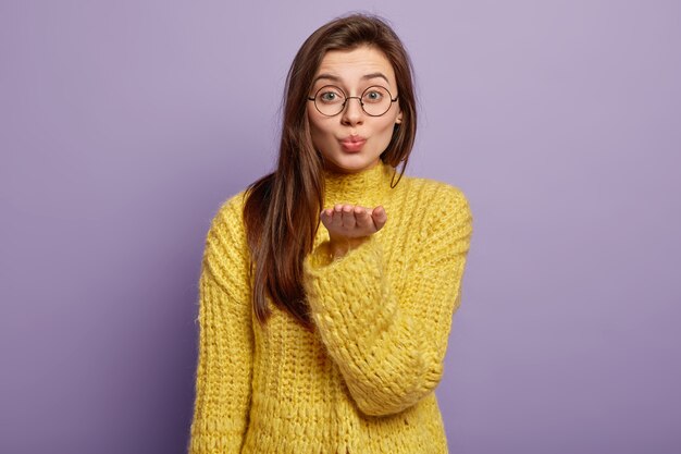 Portrait of lovely dark hared female makes air kiss, sends on distance, expresses love, stretches palm forward, wears glasses winter yellow jumper stands in  over lilac wall Body language