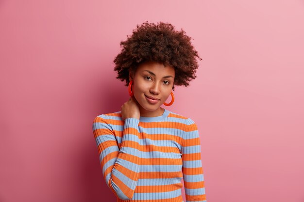 Portrait of lovely dark skinned millennial girl touches neck and looks with charming expression , sees something she wants, has curly bushy hair, dressed casually, isolated on pink wall