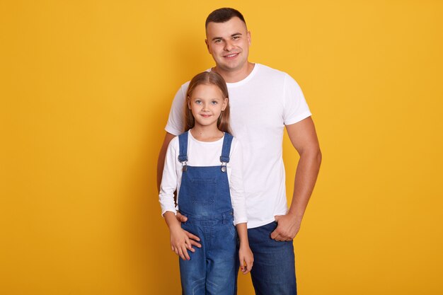 Portrait of lovely daughter smiling and standing with her handsome father isolated over yellow, family wearing casual clothing