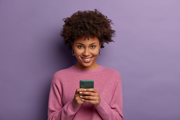 Free photo portrait of lovely ethnic woman holds modern mobile phone, uses electronic device on surfing web, looks positively , connected to wireless internet, wears casual sweater, poses indoor