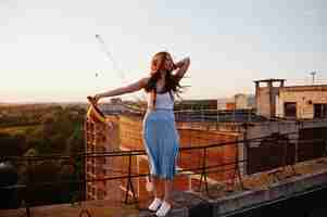 Foto gratuita ritratto di una bella ragazza in abbigliamento casual in posa sul tetto con il cappello in mano al tramonto