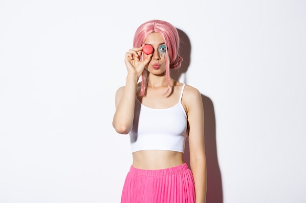 Free photo portrait of lovely girl in pink wig, dressed up for party or holiday celebration, holding delicious macaroon and pouting silly, standing.