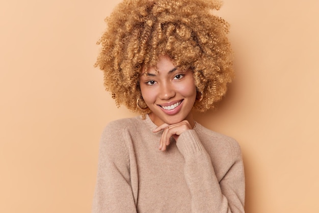 Portrait of lovely good looking curly haired young woman keeps hand under chin smiles pleasantly has glad expression dressed in casual jumper isolated over beige background. Authentic emotions