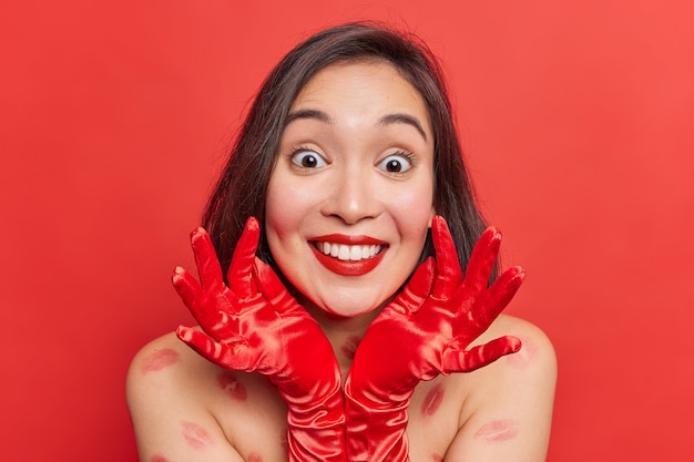 Portrait of lovely happy suprised Asian woman with dark hair wears gloves keeps hands near face wears minimal makeup stands bare shoulders indoor has kiss traces on body isolated over red wall