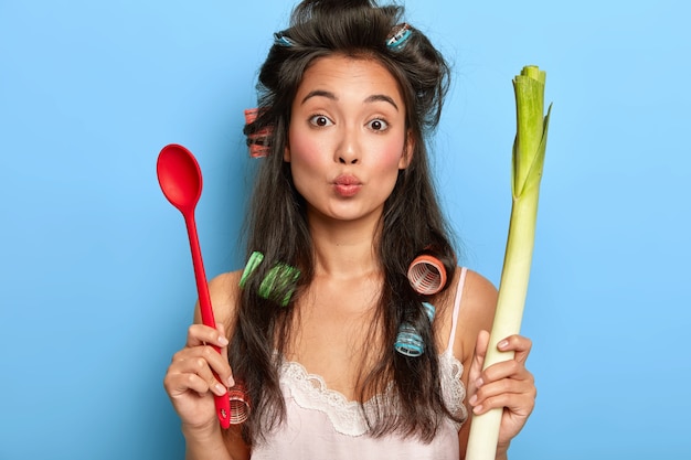 Free photo portrait of lovely housewife keeps lips rounded, wants to kiss husband, holds spoon and green fresh vegetable