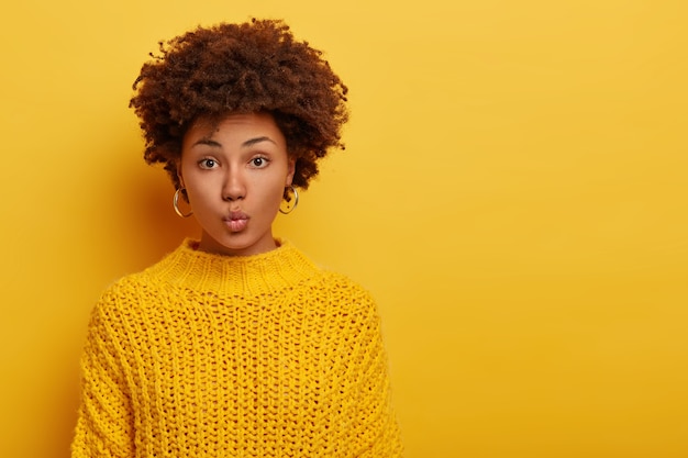 Ritratto di bella signora con acconciatura afro, mantiene le labbra piegate, indossa orecchini d'argento e maglione giallo lavorato a maglia ha lo sguardo diretto verso la telecamera si trova al coperto contro lo spazio della copia sfondo luminoso