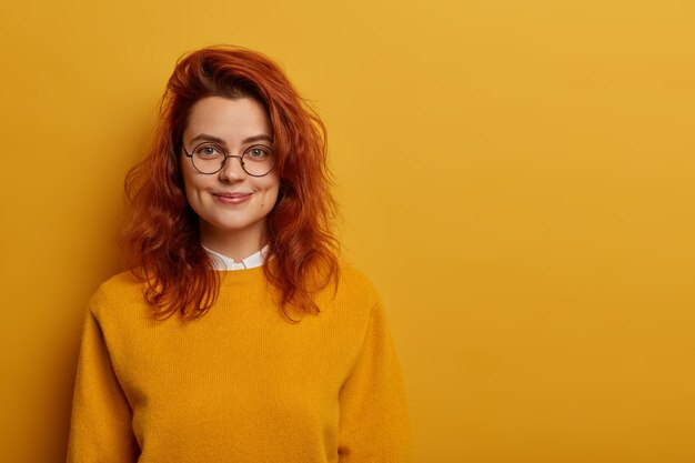 Portrait of lovely redhead woman has gentle smile, healthy skin, wears optical glasses, yellow sweater, enjoys day off