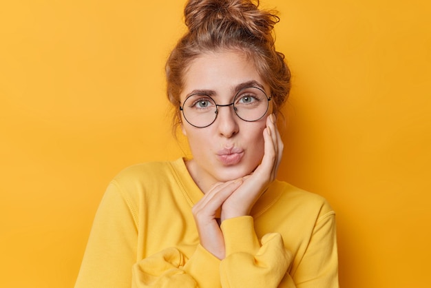 Portrait of lovely tender young woman has folded lips keeps hand on cheek looks gently at camera wears round transparent glasses and jumper poses against yellow background has romantic expression