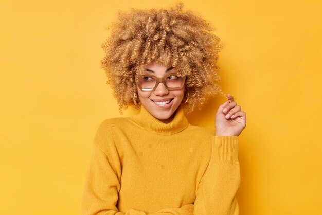 Portrait of lovely thoughtful woman with happy dreamy expression bites lips and looks away has good mood wears spectacles and warm sweater isolated over vivid yellow background Positive emotions