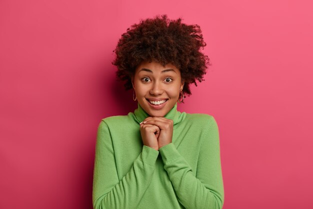 Portrait of lovely woman has curly hairstyle, looks gladfully at camera, holds hands under chin, wears casual green poloneck, has cute smile, white teeth